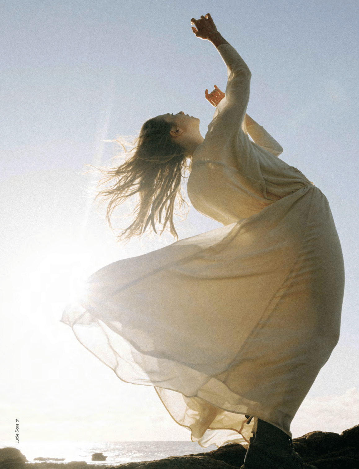 Woman in a white dress standing on a beach with arms raised, sunlit background