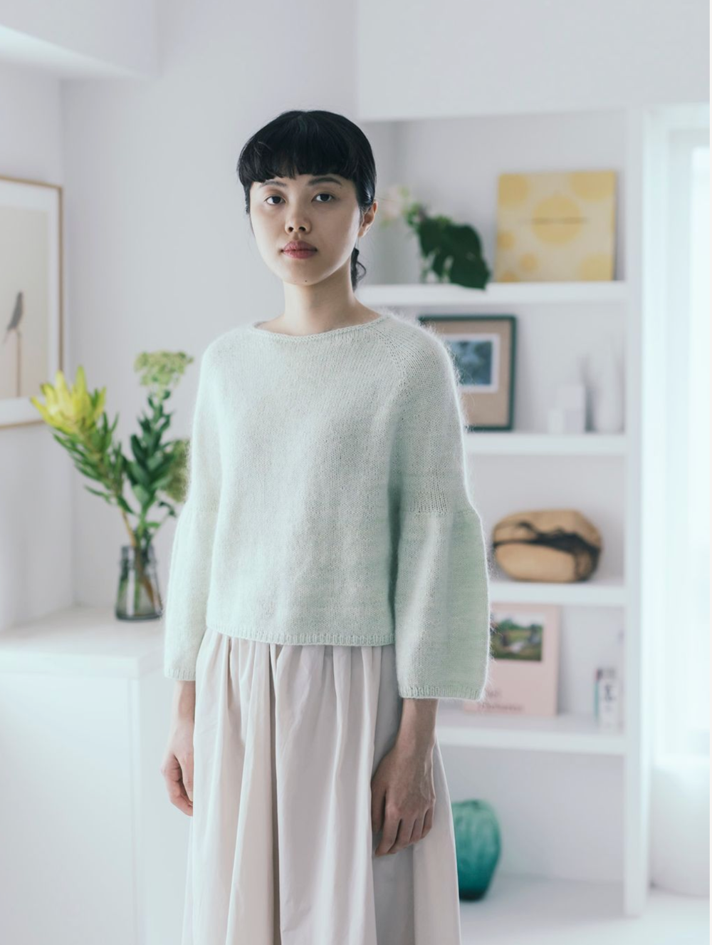 Person wearing a light green sweater and beige pants in a bright room with plants and books.