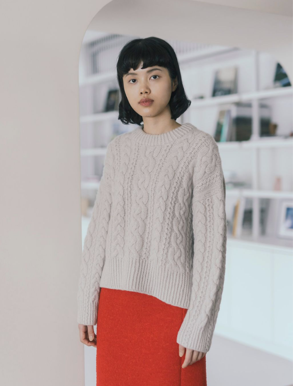 Woman wearing a beige cable knit sweater and red pants in an indoor setting.
