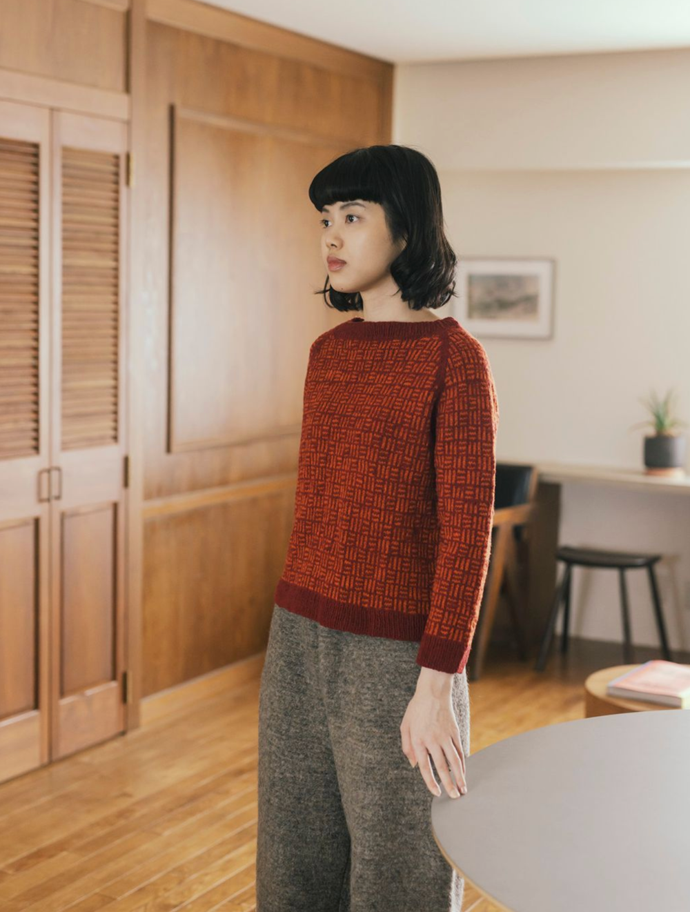 woman wearing a graphic red and orange motif sweater against a background of white wall and wooden panelling.
