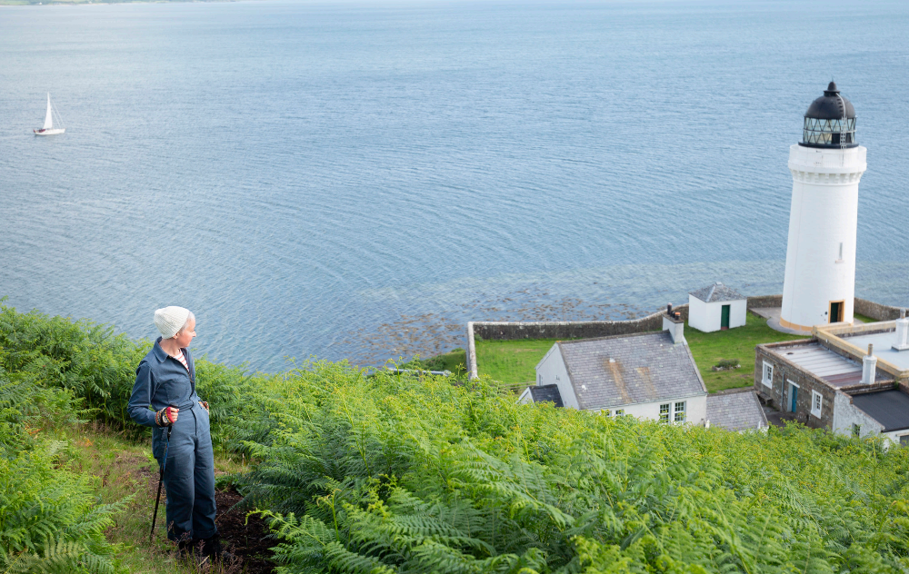 Person with a walking stick standing on a grassy hill overlooking a lighthouse and body of water.