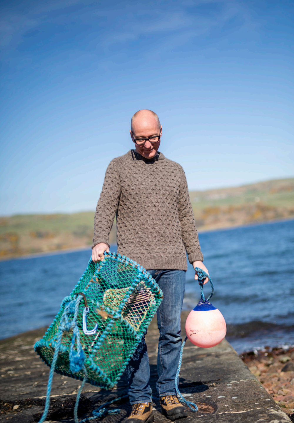 Man holding a green lobster trap and pink buoy by a body of water.