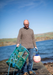 Man holding a green lobster trap and pink buoy by a body of water.