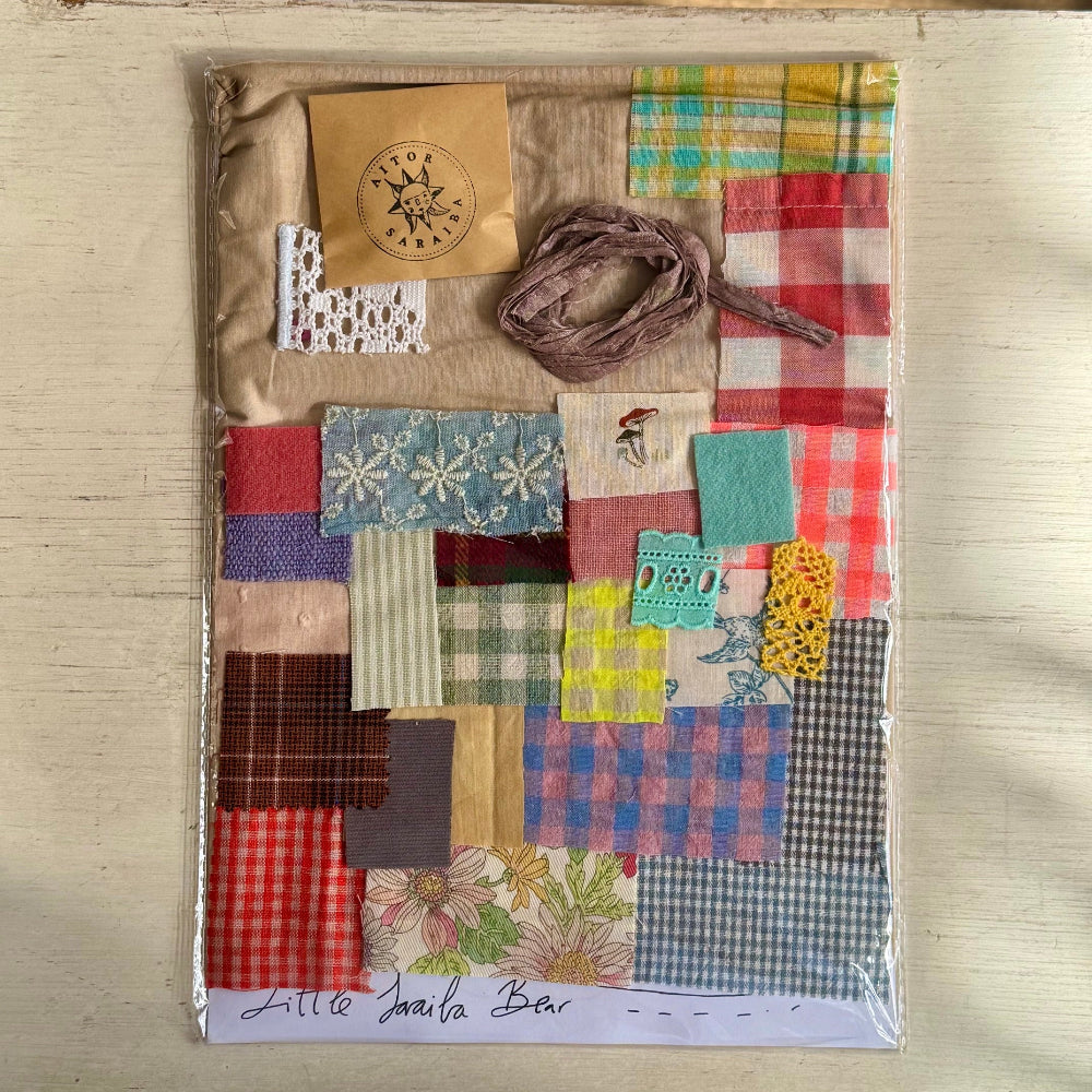 A handmade patchwork fabric kit in a clear plastic sleeve, featuring various colorful fabric squares, plaid patterns, lace, and a brown ribbon on a light wood background.