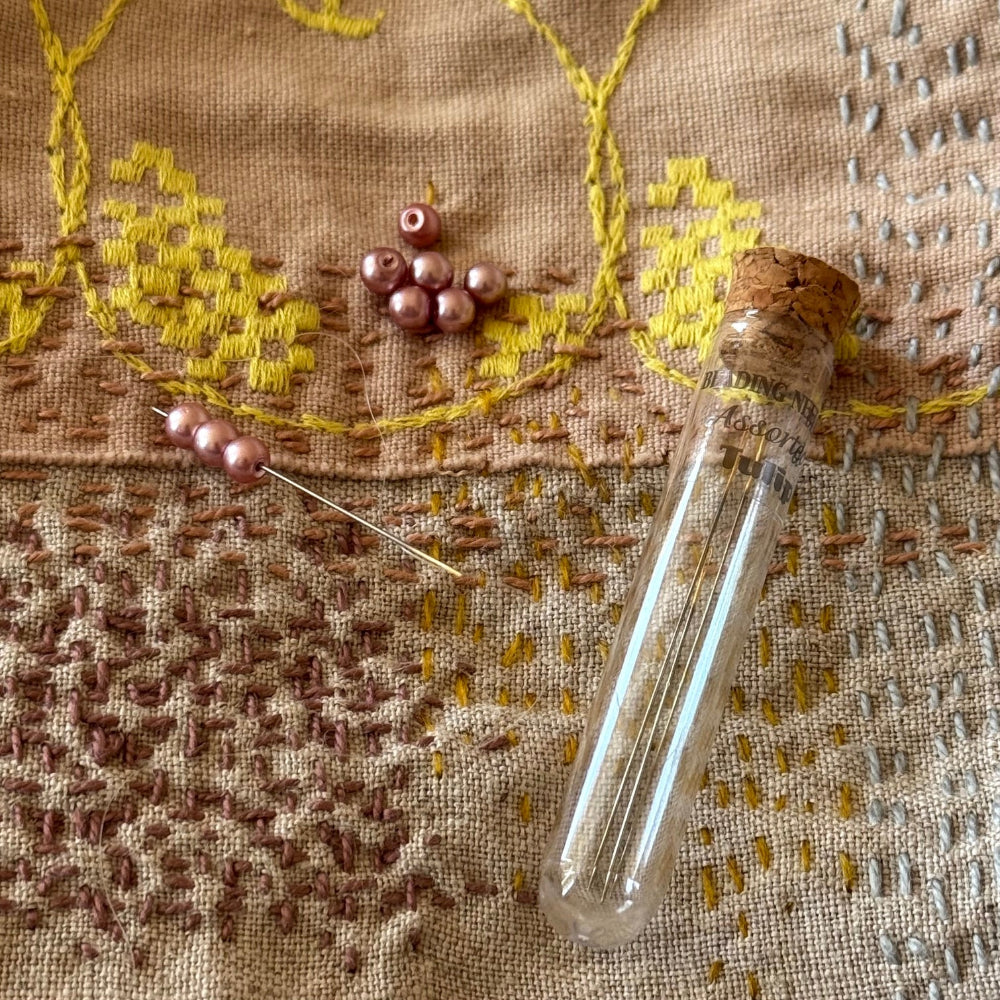 A glass vial with a cork stopper and a needle threaded with pink beads, arranged on a textured beige fabric with yellow and brown embroidered patterns.