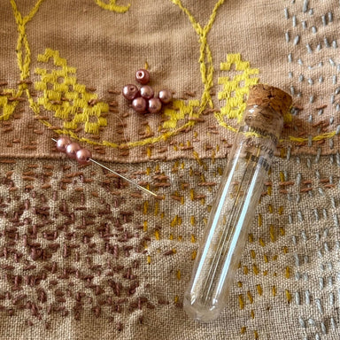 A glass vial with a cork stopper and a needle threaded with pink beads, arranged on a textured beige fabric with yellow and brown embroidered patterns.
