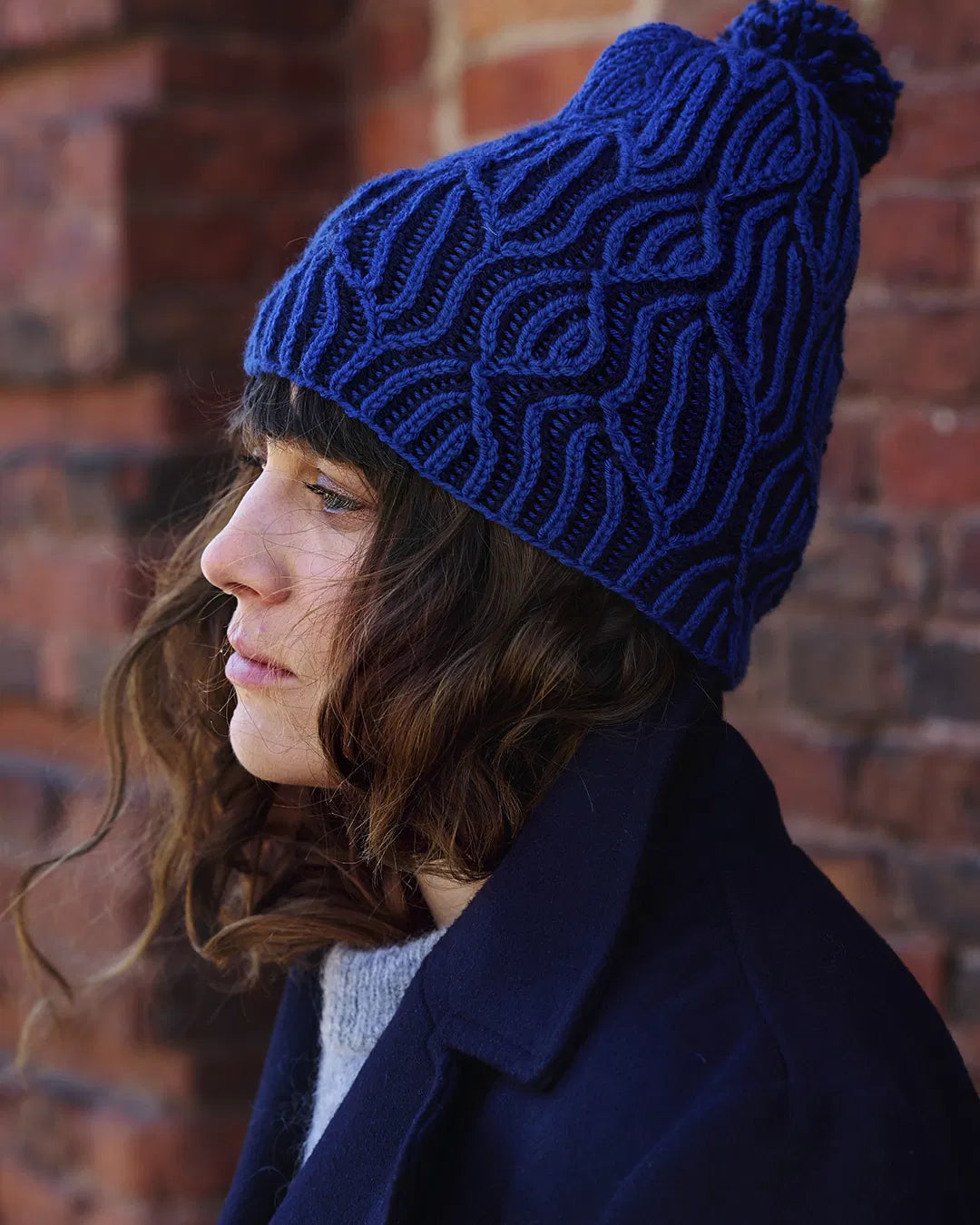 Person wearing a blue knitted beanie with a pom-pom against a brick wall.