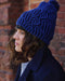 Person wearing a blue knitted beanie with a pom-pom against a brick wall.