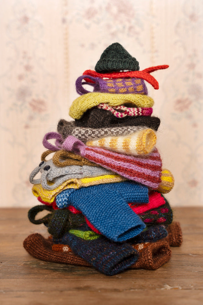 A stack of colorful handmade knit baby socks and booties arranged in a pyramid shape on a wooden surface, with a soft floral wallpaper background.