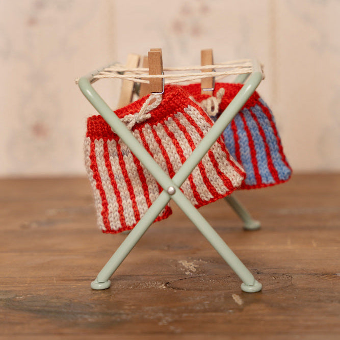 A miniature light green metal folding clothes rack holding two small knitted socks, one red and white striped and one blue and white striped, with wooden clothespins on a rustic wooden tabletop against a soft floral background.