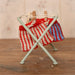 A miniature light green metal folding clothes rack holding two small knitted socks, one red and white striped and one blue and white striped, with wooden clothespins on a rustic wooden tabletop against a soft floral background.