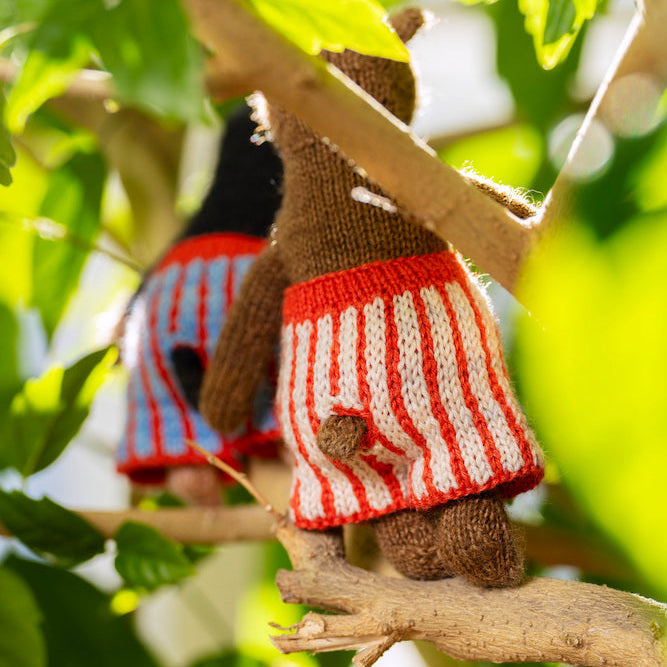 A close-up of a hand-knit brown teddy bear wearing a red and white striped sweater vest, perched on a tree branch surrounded by green leaves.