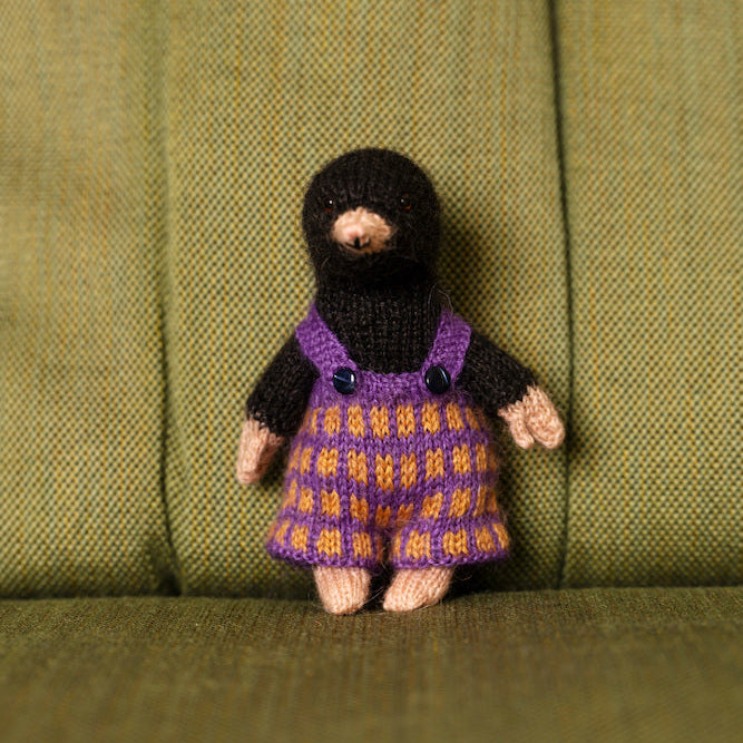 A small handmade knitted doll with a black balaclava-style head and purple-and-orange patterned dress, standing against an olive green upholstered chair.