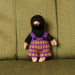 A small handmade knitted doll with a black balaclava-style head and purple-and-orange patterned dress, standing against an olive green upholstered chair.
