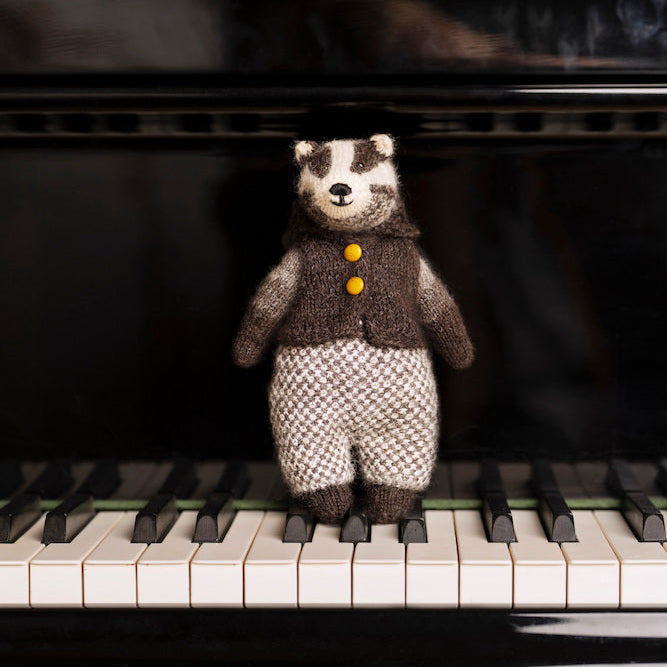 A small knitted badger toy wearing a brown vest with yellow buttons and patterned trousers, standing on the piano keys in front of a black grand piano.