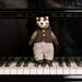 A small knitted badger toy wearing a brown vest with yellow buttons and patterned trousers, standing on the piano keys in front of a black grand piano.