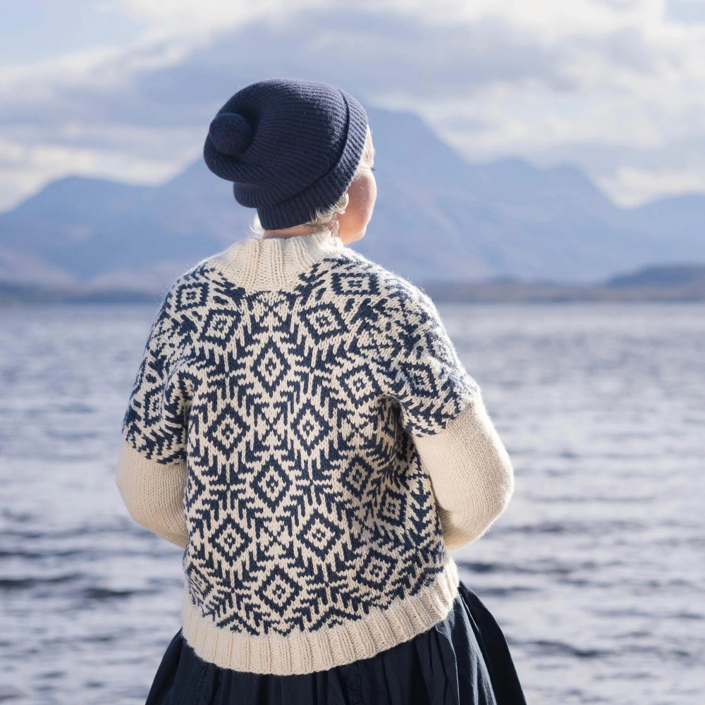 Person wearing a patterned sweater and dark hat standing by a lake with mountains in the background