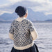 Person wearing a patterned sweater and dark hat standing by a lake with mountains in the background