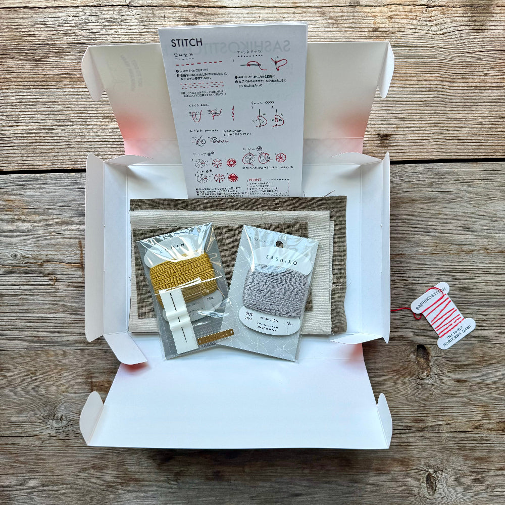 A Sashiko stitch embroidery kit in a white cardboard box with a window showing thread spools inside, placed on a wooden surface. A red thread spool tag hangs to the right with the text 'SASHIKOSTITCH HORIZOWA NAMI'.