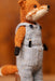 A close-up of a knitted fox doll wearing light gray overalls with black buttons, standing against a warm brown blurred background.