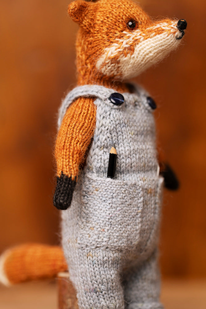 A close-up of a knitted fox doll wearing light gray overalls with black buttons, standing against a warm brown blurred background.