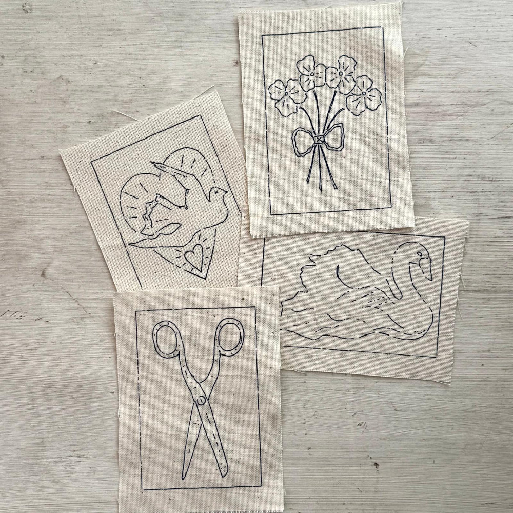 Four small square fabric patches with black line drawings laid on a light wood surface. The designs show a dove in a heart, a bouquet of flowers tied with a bow, a swan, and a pair of scissors.