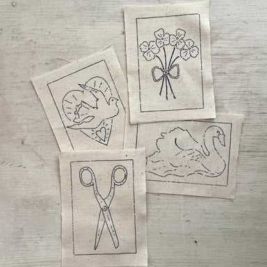 Four small square fabric patches with black line drawings laid on a light wood surface. The designs show a dove in a heart, a bouquet of flowers tied with a bow, a swan, and a pair of scissors.
