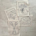 Four small square fabric patches with black line drawings laid on a light wood surface. The designs show a dove in a heart, a bouquet of flowers tied with a bow, a swan, and a pair of scissors.