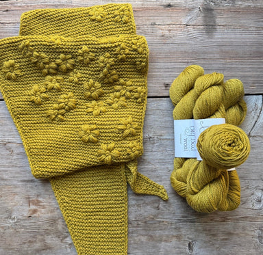 A mustard yellow knitted scarf with raised flower patterns laid flat on a rustic wooden surface, next to a skein of matching yarn with a white label that reads 'PooLoo Organic'.