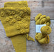A mustard yellow knitted scarf with raised flower patterns laid flat on a rustic wooden surface, next to a skein of matching yarn with a white label that reads 'PooLoo Organic'.
