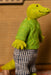 A knitted crocodile doll wearing a bright green sweater and blue-and-white striped trousers, standing indoors against a warm brown textured background.