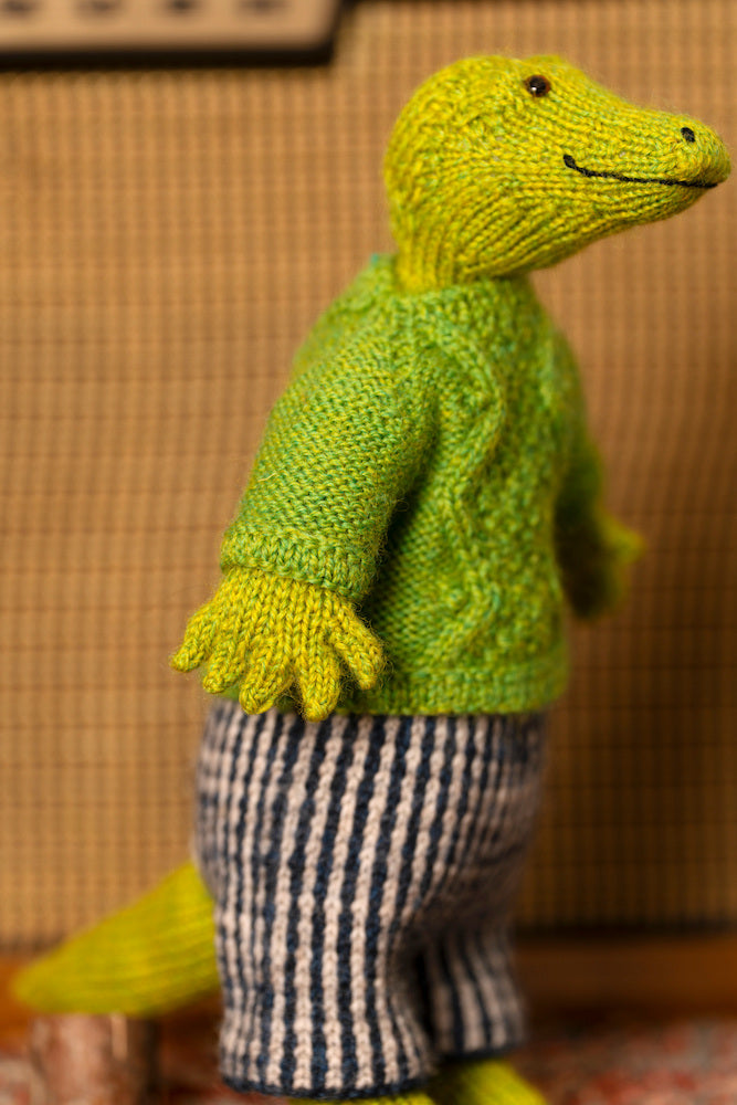 A knitted crocodile doll wearing a bright green sweater and blue-and-white striped trousers, standing indoors against a warm brown textured background.