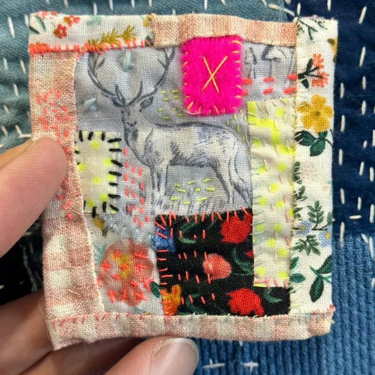 Small embroidered patch with floral and deer design held by a hand on a textured blue fabric background.