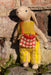 A knitted bunny doll with long ears stands outdoors against a rustic stone wall. The bunny is dressed in yellow knit clothing with a red-and-white checkered bag hanging from its neck.