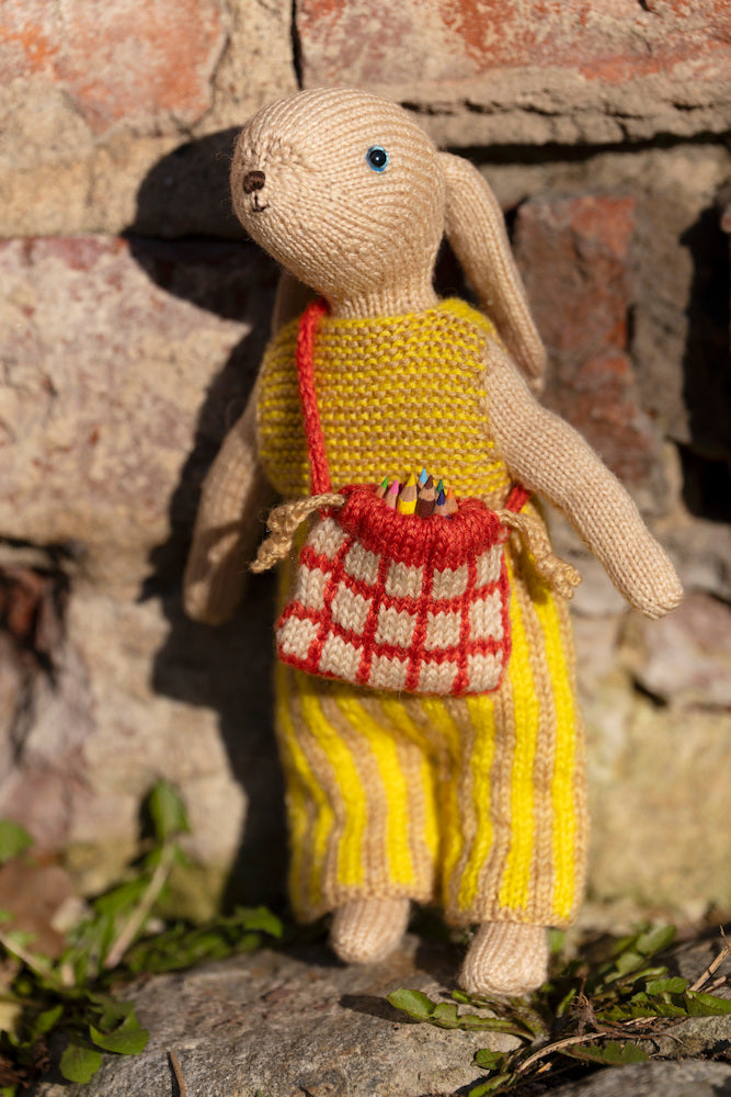 A knitted bunny doll with long ears stands outdoors against a rustic stone wall. The bunny is dressed in yellow knit clothing with a red-and-white checkered bag hanging from its neck.