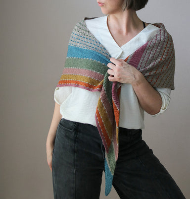 Person wearing a colorful striped shawl over a white shirt and dark pants on a neutral background