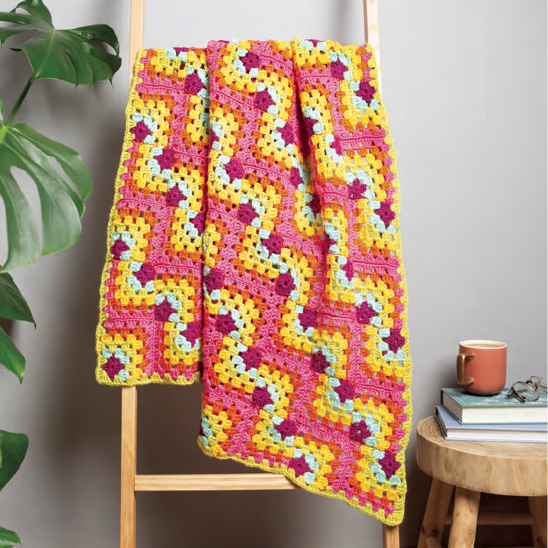 Colorful crocheted blanket draped over a wooden ladder against a neutral wall.