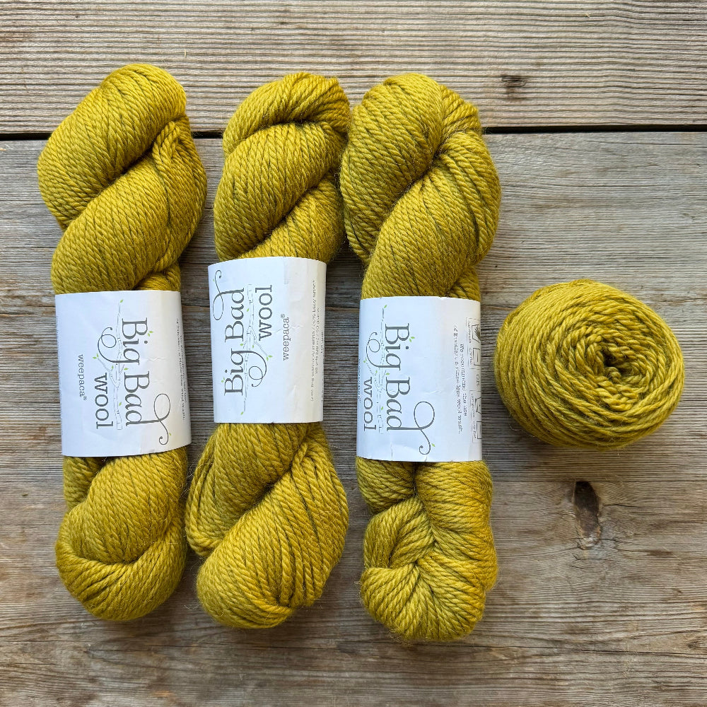 Three skeins of mustard yellow wool yarn and one matching ball of yarn with white paper labels reading Big Bad Wool, arranged on a rustic wooden surface.