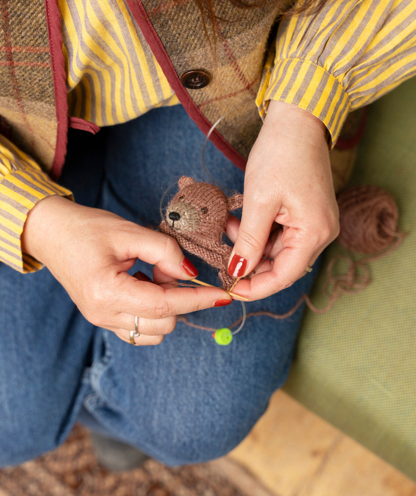 A close-up of a person's hands knitting a small brown teddy bear with knitting needles, wearing a yellow and brown striped shirt, a tweed vest, and blue jeans.
