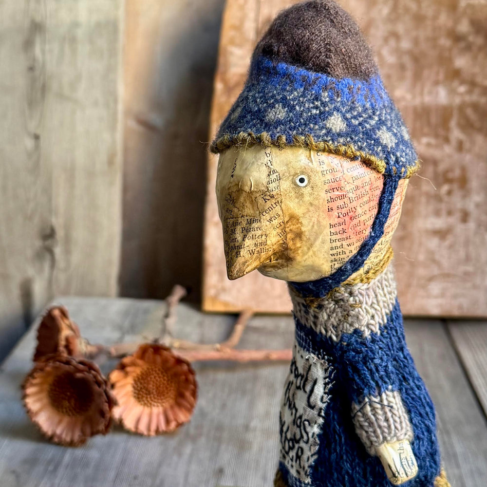 A rustic handmade doll figurine with a paper-textured face and body, wearing a blue and cream knit hat and scarf, styled on a wooden surface with dried acorns.