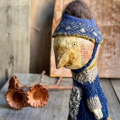 A rustic handmade doll figurine with a paper-textured face and body, wearing a blue and cream knit hat and scarf, styled on a wooden surface with dried acorns.