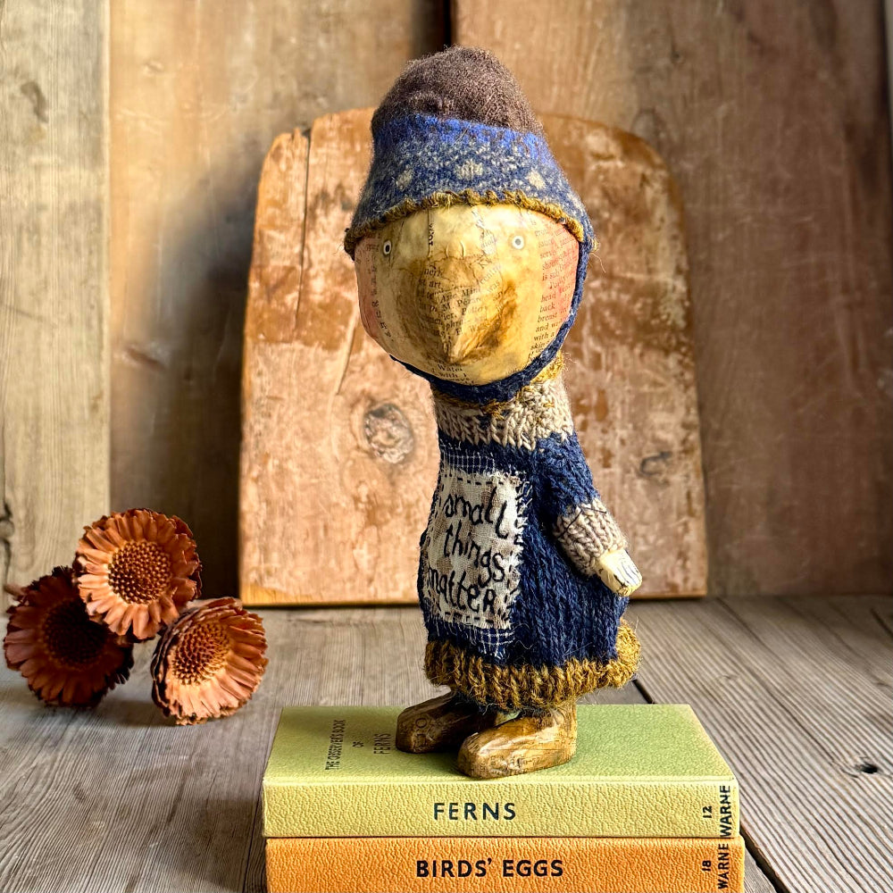 A rustic handmade wooden bird figurine wearing a blue and brown knit hat and sweater, standing on two vintage books with a wooden cutting board and dried flowers in the background.