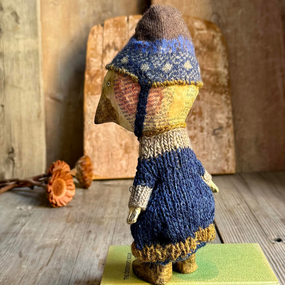 A handmade knitted bird doll shown from the back, wearing a blue patterned hat and a blue dress with cream and mustard accents, standing on a green surface against a rustic wooden background.