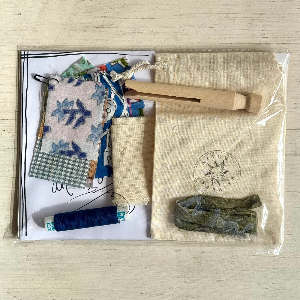 A packaged sewing or embroidery kit laid flat on a light wood surface, including patterned fabric pieces, a wooden needle holder, a beige fabric pouch with a printed compass-style logo, a blue spool of thread, and a small olive-green fabric piece inside clear plastic wrapping.