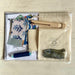 A packaged sewing or embroidery kit laid flat on a light wood surface, including patterned fabric pieces, a wooden needle holder, a beige fabric pouch with a printed compass-style logo, a blue spool of thread, and a small olive-green fabric piece inside clear plastic wrapping.