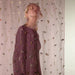 A woman wearing a long-sleeved mauve dress with gold and black embroidered details, standing in front of a sheer curtain decorated with small gold and black embellishments.