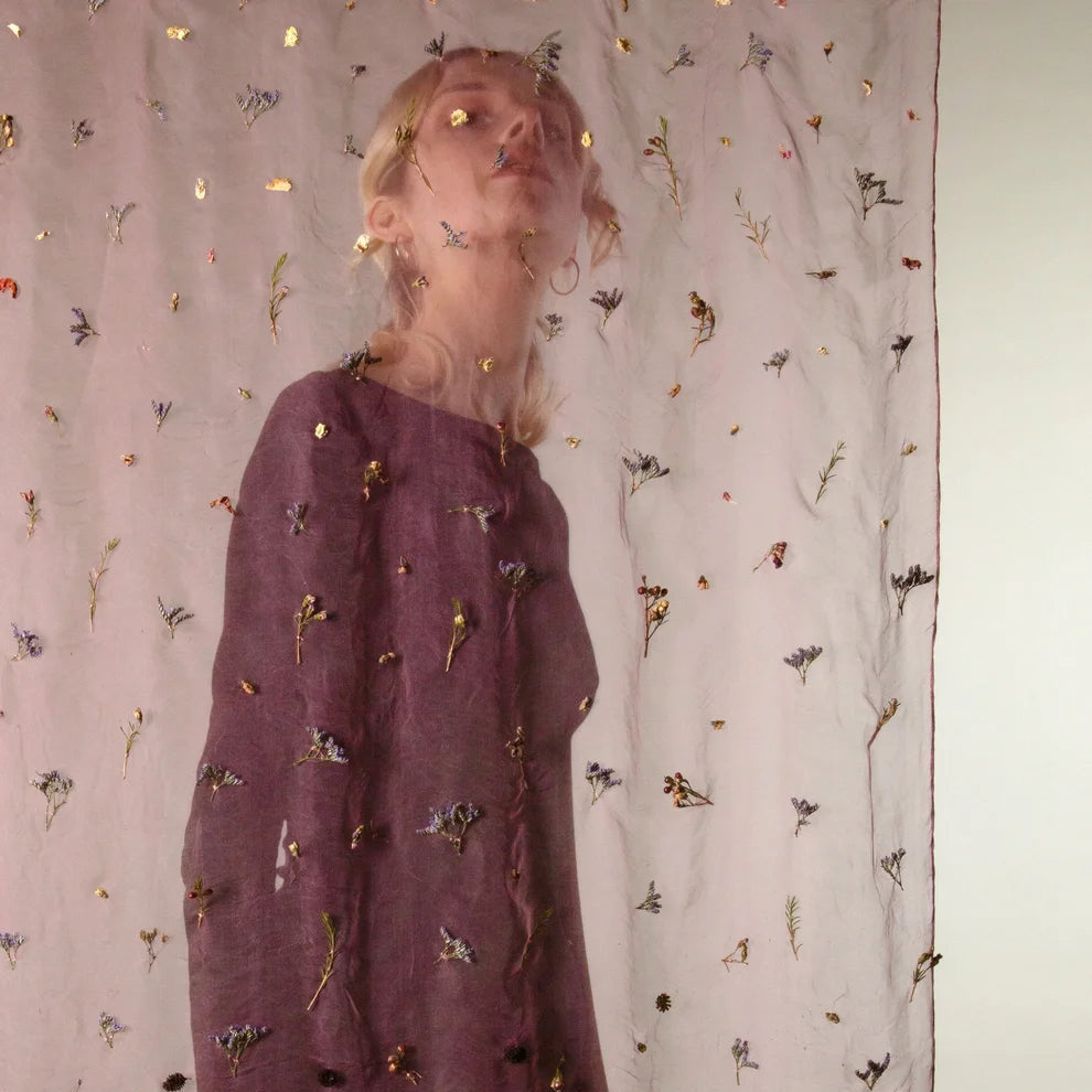 A woman wearing a long-sleeved mauve dress with gold and black embroidered details, standing in front of a sheer curtain decorated with small gold and black embellishments.