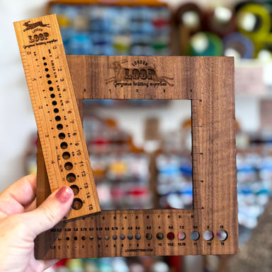 Wooden knitting needle gauge held in front of a blurred store shelf.