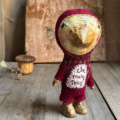 A handmade doll or figurine wearing a knitted burgundy hooded outfit with a paper face and gold shoes, standing on a rustic wooden surface with a small acorn prop in the background.