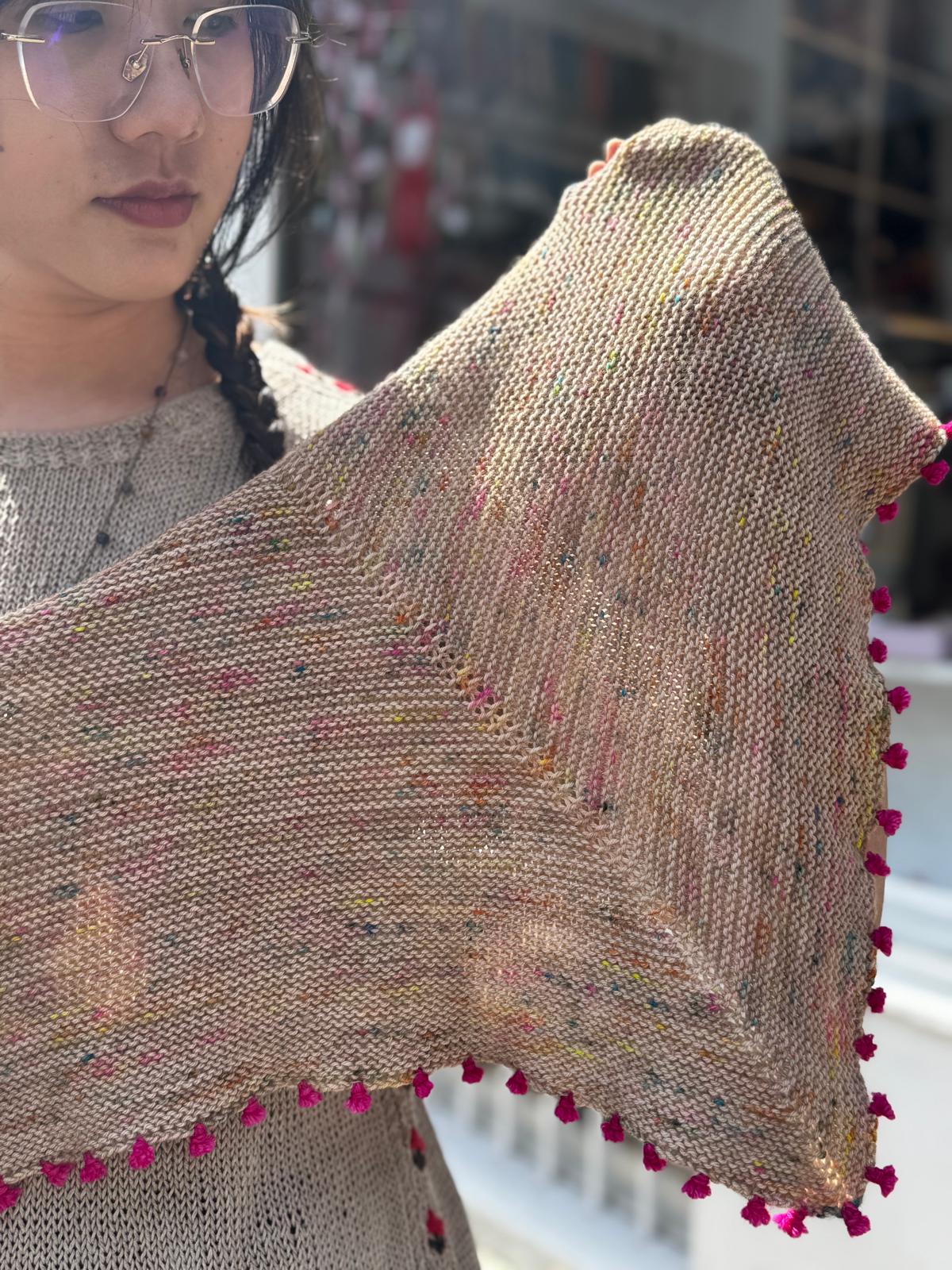 A person wearing glasses holds up a chunky knit triangular shawl in a warm beige/tan color with multicolored flecks and bright pink pom-pom trim along the edges.